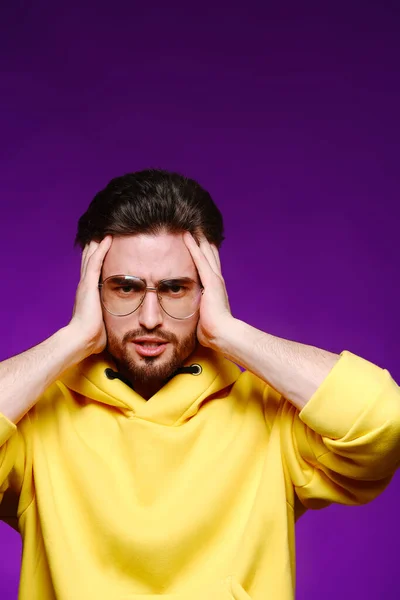 Un jeune homme de 25-30 ans dans des lunettes et un sweat-shirt jaune pose émotionnellement tenant sa tête sur un fond violet. — Photo