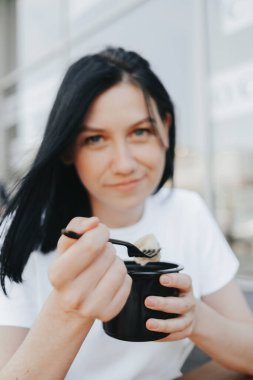 Beyaz tişörtlü, siyah saçlı genç bir kadın bir kafedeki açık bir verandada oturup tek kullanımlık plastik tabaklardan yemek yiyor.. 