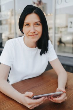 Beyaz tişörtlü, siyah saçlı genç bir kadın bir kafedeki açık bir verandada oturuyor ve siparişini beklerken akıllı telefon kullanıyor..