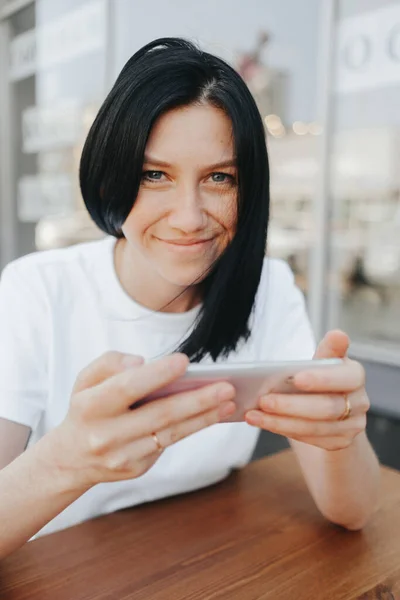 En ung kvinna med svart hår i vit T-shirt sitter på en öppen veranda på ett café och använder en smartphone medan hon väntar på sin beställning och hämtmat. — Stockfoto