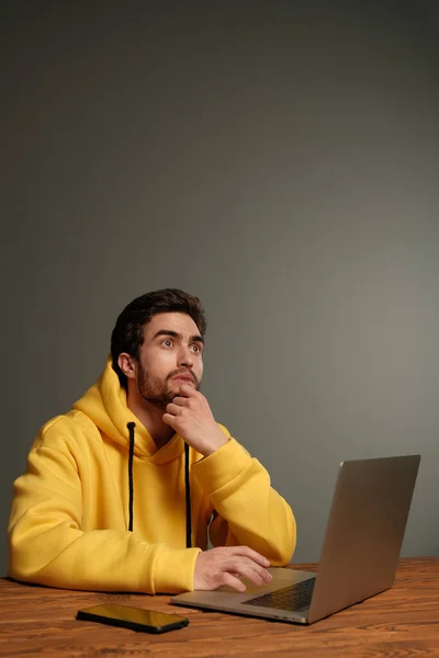 Un joven con diferentes emociones en una sudadera amarilla funciona con un ordenador portátil y un teléfono inteligente . —  Fotos de Stock
