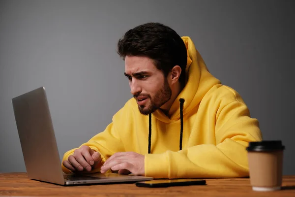 A young guy with different emotions in a yellow sweatshirt works using a laptop and smartphone.
