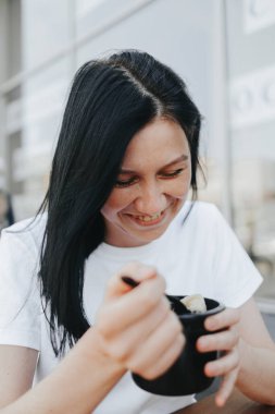 Beyaz tişörtlü, siyah saçlı genç bir kadın bir kafedeki açık bir verandada oturup tek kullanımlık plastik tabaklardan yemek yiyor.. 