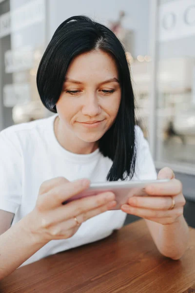 Młoda kobieta z czarnymi włosami w białej koszulce siedzi na otwartej werandzie w kawiarni i używa smartfona czekając na zamówienie i jedzenie na wynos.. — Zdjęcie stockowe