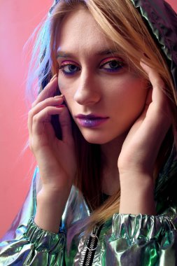 Parlak makyajlı güzel bir kız metalik bir rüzgarlıkla moda pembe neon arka planda poz veriyor..
