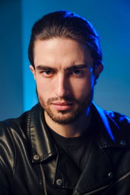25-30 yaşlarında deri ceketli genç bir adam mavi arka planda poz veriyor..