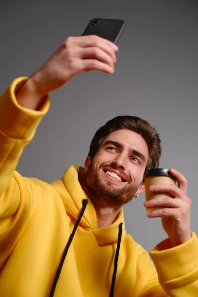 Joven chico sonriente en una sudadera amarilla con un vaso de papel desechable de café en la mano hace selfie en un teléfono inteligente —  Fotos de Stock