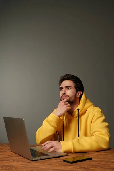 Un joven con diferentes emociones en una sudadera amarilla funciona con un ordenador portátil y un teléfono inteligente . —  Fotos de Stock