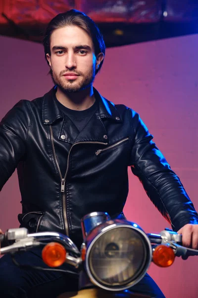 Um jovem de 25-30 anos em uma jaqueta de couro e jeans preto posando enquanto sentado em um ciclomotor em um fundo roxo . — Fotografia de Stock