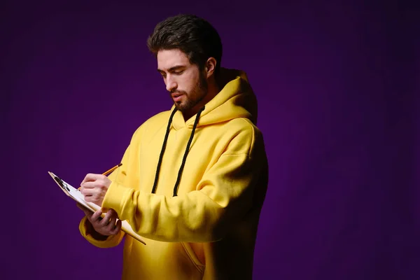 A young man of 25-30 years in glasses and a yellow sweatshirt holding a clipboard with clip in one hand and a pencil in the other — Stock Photo, Image