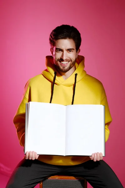 Un jeune homme de 25 à 30 ans en sweat-shirt jaune assis sur une vieille colonne de musique sur fond de mur rose et tenant un livre ou un album ouvert. — Photo