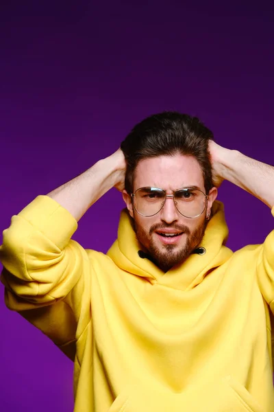 Un jeune homme de 25-30 ans dans des lunettes et un sweat-shirt jaune pose émotionnellement tenant sa tête sur un fond violet. — Photo
