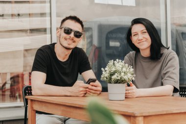 Genç bir kadın ve erkek bir kafede açık bir verandada oturur ve sipariş ve paket beklerken akıllı telefon kullanır.. 