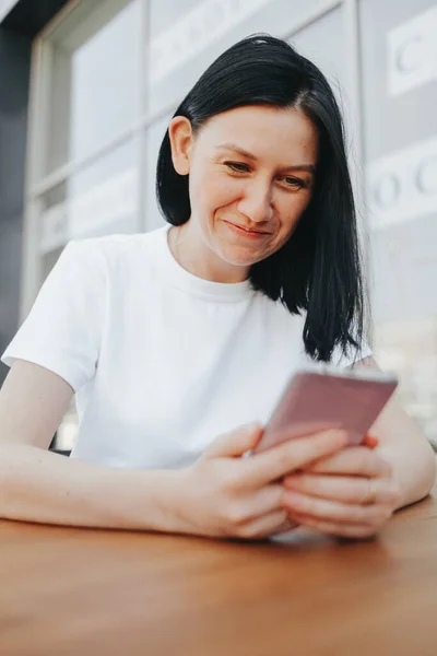 Młoda kobieta z czarnymi włosami w białej koszulce siedzi na otwartej werandzie w kawiarni i używa smartfona czekając na zamówienie i jedzenie na wynos.. — Zdjęcie stockowe