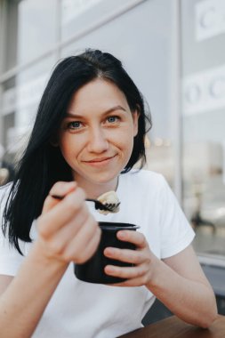 Beyaz tişörtlü, siyah saçlı genç bir kadın bir kafedeki açık bir verandada oturup tek kullanımlık plastik tabaklardan yemek yiyor.. 