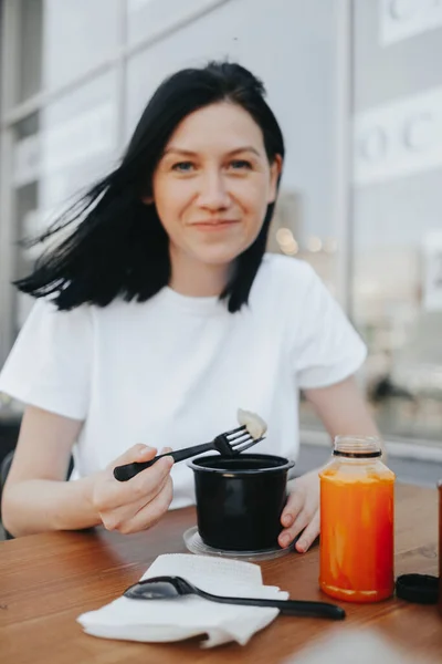 Молода жінка з чорним волоссям у білій футболці сидить на відкритій веранді в кафе і їсть їжу з одноразового пластикового посуду . — стокове фото