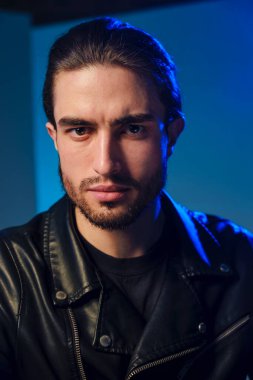 25-30 yaşlarında deri ceketli genç bir adam mavi arka planda poz veriyor..