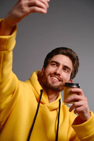 Jeune homme souriant dans un sweat-shirt jaune avec un verre de papier jetable de café à la main fait selfie sur un smartphone — Photo