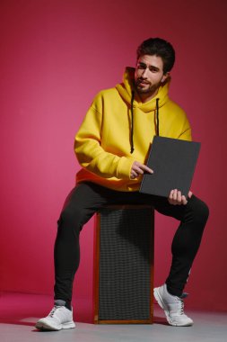 25-30 yaşlarında, sarı kazaklı, pembe duvarlı eski bir müzik sütunu üzerinde oturan ve elinde kapalı bir kitap ya da albüm tutan genç bir adam.. 