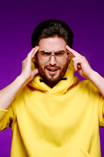 Un jeune homme de 25-30 ans dans des lunettes et un sweat-shirt jaune pose émotionnellement tenant sa tête sur un fond violet. — Photo