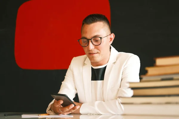 Un joven profesor de escuela, un hombre con una chaqueta blanca y gafas utiliza un teléfono inteligente y se está preparando para una nueva lección . —  Fotos de Stock