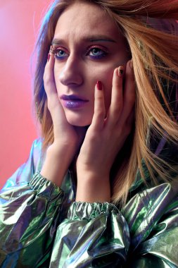 Parlak makyajlı güzel bir kız metalik bir rüzgarlıkla moda pembe neon arka planda poz veriyor..