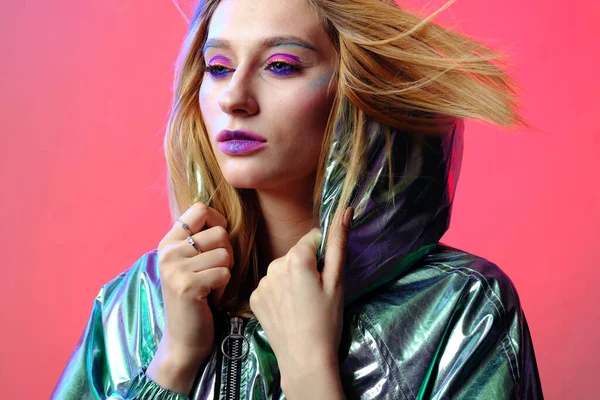 Parlak makyajlı güzel bir kız metalik bir rüzgarlıkla moda pembe neon arka planda poz veriyor..