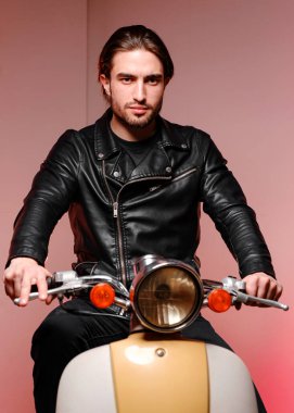 25-30 yaşlarında deri ceketli ve siyah kotlu genç bir adam mor bir arka planda motosikletin üzerinde otururken poz veriyor..