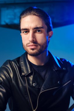 25-30 yaşlarında deri ceketli genç bir adam mavi arka planda poz veriyor..