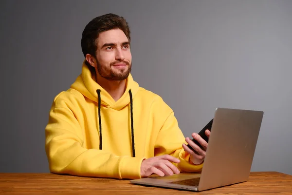 Un joven con diferentes emociones en una sudadera amarilla funciona con un ordenador portátil y un teléfono inteligente . —  Fotos de Stock