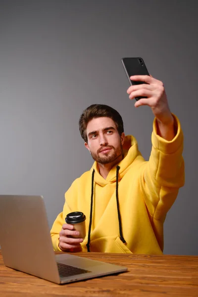 Junger lächelnder Kerl im gelben Sweatshirt mit einem Einwegpapierglas Kaffee in der Hand macht Selfie auf dem Smartphone — Stockfoto