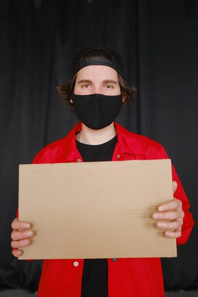 Un joven de 25-30 años con una máscara protectora negra, gafas amarillas, una gorra y una chaqueta roja sostiene un cartel de cartón vacío en sus manos . — Foto de Stock