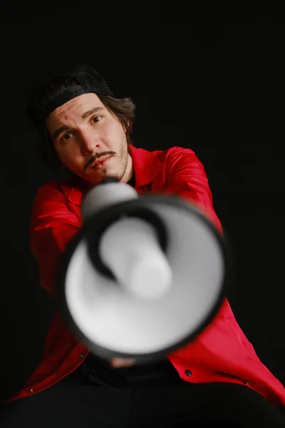 Эмоциональный молодой человек 25-30 лет в черной кепке и в красной куртке сидит на синей бочке и держит мегафон в руках на темном фоне. Концепция протеста — стоковое фото