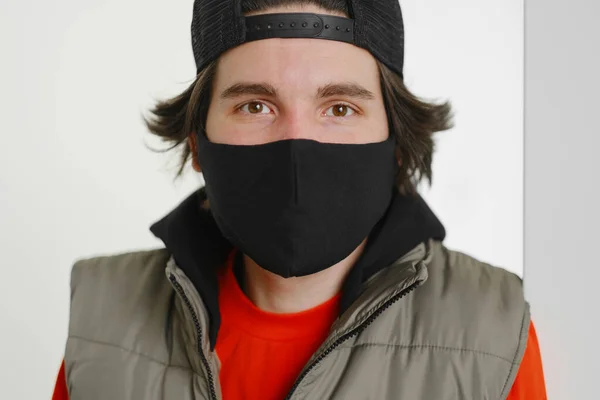 Un jeune homme de 25 à 30 ans portant un masque de protection noir, une casquette et une veste rouge posant sur un fond gris. — Photo