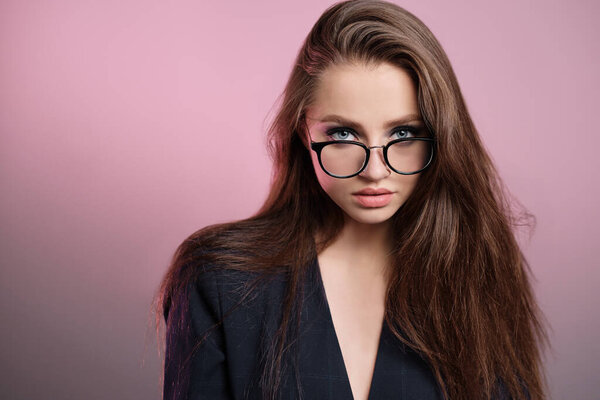A young girl 20-25 years old in glasses, a jacket in the image of a teacher posing on a pink background. 