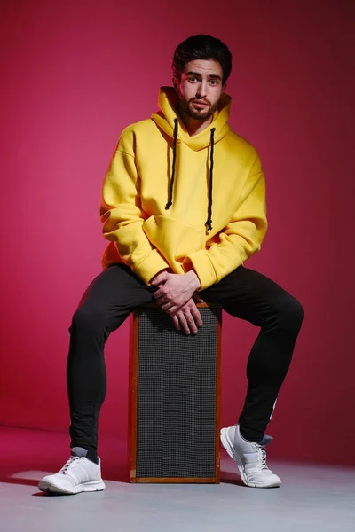Mladý muž 25-30 let ve žluté mikině sedící na starém hudebním sloupku na růžovém pozadí a ukazující různé emoce. — Stock fotografie