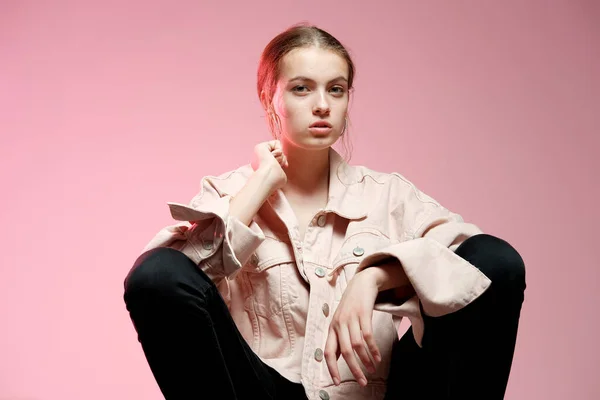 Una chica en un vaquero rosa en jeans negros se sienta en un taburete de madera blanca y posa sobre un fondo rosa . — Foto de Stock