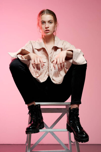 A girl in a pink jeans in black jeans sits on a white wooden bar stool and poses on a pink background.