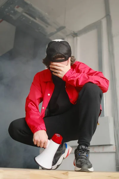 Joven emocional de 25-30 años con una máscara protectora negra, una gorra y una chaqueta roja sosteniendo un megáfono sobre un fondo oscuro . — Foto de Stock