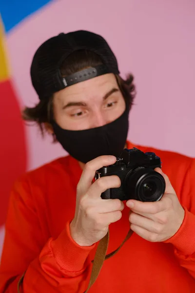 Photographe masculin. Un jeune homme de 25 à 30 ans portant un masque de protection noir et un sweat-shirt rouge tient dans ses mains un appareil photo sans miroir. — Photo