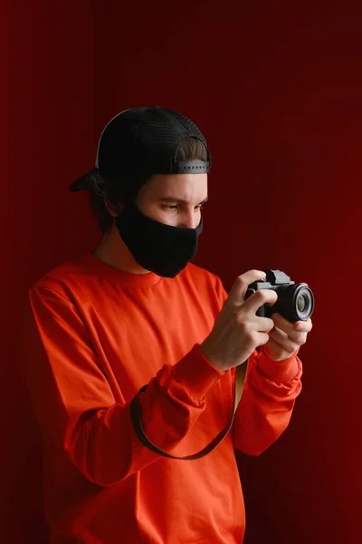 Fotografo maschio. Un giovane uomo di 25-30 anni con una maschera protettiva nera e una felpa rossa tiene in mano una macchina fotografica senza specchio. — Foto Stock