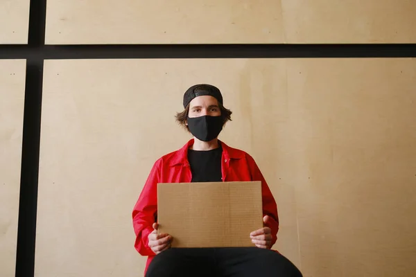 Un joven de 25-30 años con una máscara protectora negra, gafas amarillas, una gorra y una chaqueta roja sostiene un cartel de cartón vacío en sus manos . — Foto de Stock