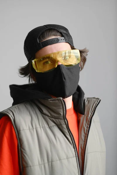 Молодой человек 25-30 лет в черной защитной маске, желтых очках, кепке и красной куртке позирует на сером фоне. — стоковое фото