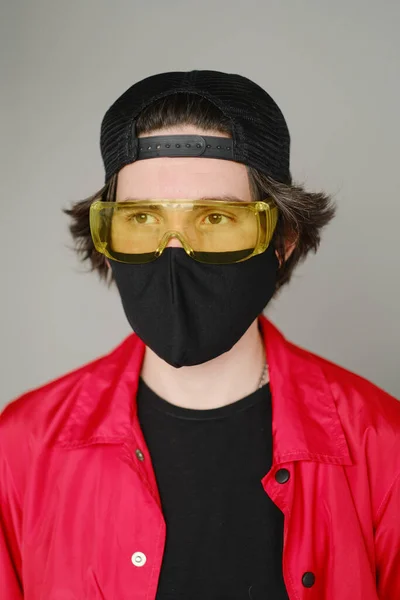 Ein junger Mann von 25-30 Jahren mit schwarzer Schutzmaske, gelber Brille, Mütze und roter Jacke posiert vor grauem Hintergrund. — Stockfoto