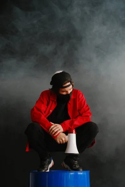 Эмоциональный молодой человек 25-30 лет в черной защитной маске, кепке и красной куртке держит мегафон на темном фоне. — стоковое фото