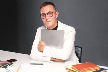 30-35 yaşlarında, beyaz ceketli, siyah balıkçı yakalı, gözlüklü ve kısa saçlı genç bir adam elinde boş bir tabela tutuyor.