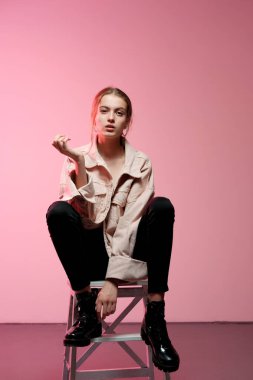 Siyah kotlu pembe bir kız beyaz tahta bir bar taburesinde oturur ve pembe bir arka planda poz verir..