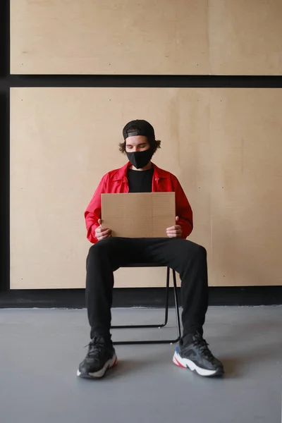 Un joven de 25-30 años con una máscara protectora negra, gafas amarillas, una gorra y una chaqueta roja sostiene un cartel de cartón vacío en sus manos . — Foto de Stock