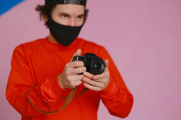 Photographe masculin. Un jeune homme de 25 à 30 ans portant un masque de protection noir et un sweat-shirt rouge tient dans ses mains un appareil photo sans miroir. — Photo