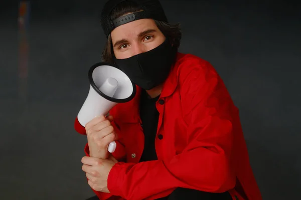 Jeune homme émotionnel de 25 à 30 ans portant un masque de protection noir, une casquette et une veste rouge tenant un mégaphone sur un fond sombre. — Photo